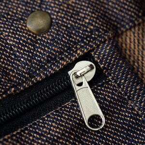 May include: Close-up of a zipper on a brown and blue fabric bag. The zipper is silver and has a black pull tab.