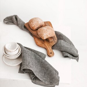May include: A rustic still life featuring two loaves of bread on a wooden cutting board, accompanied by small white bowls and a checkered gray and white linen cloth. The scene is set against a bright white background.