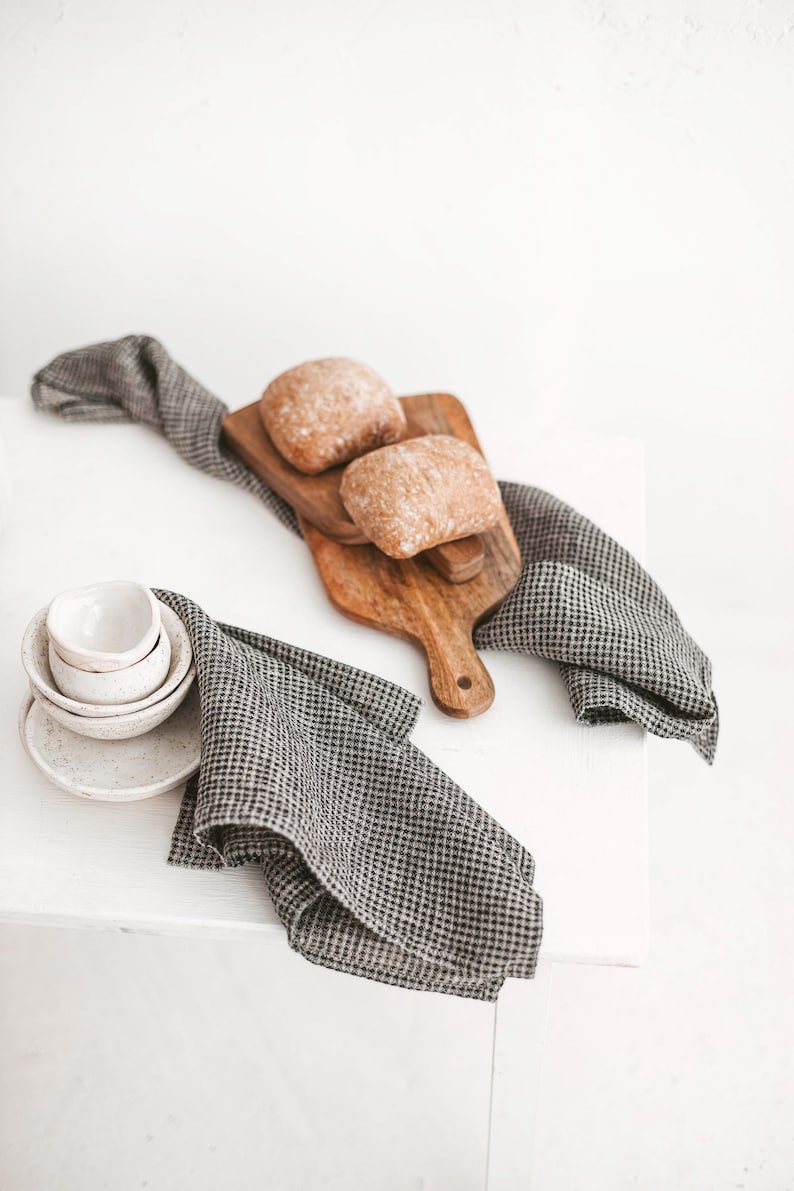 May include: A rustic still life featuring two loaves of bread on a wooden cutting board, accompanied by small white bowls and a checkered gray and white linen cloth. The scene is set against a bright white background.