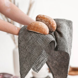 May include: Two freshly baked, golden-brown bread rolls rest on a black and white checkered linen napkin. The napkin is held up, showcasing the texture and pattern. The scene suggests a rustic, home-baked theme.