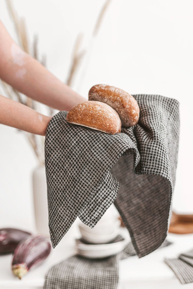 May include: Two freshly baked, golden-brown bread rolls rest on a black and white checkered linen napkin. The napkin is held up, showcasing the texture and pattern. The scene suggests a rustic, home-baked theme.