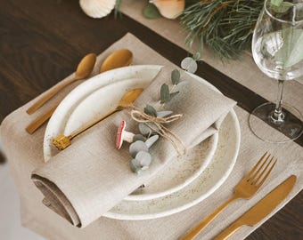 May include: A table setting with a neutral linen runner, white speckled plates, and gold-toned cutlery. A folded linen napkin is adorned with eucalyptus and a mushroom accent. Decorative elements include candles and paper ornaments.