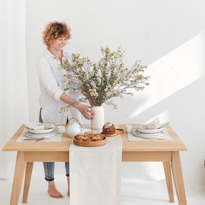 A woman putting a vase with wildflowers on a table decorated with a herri,ngbone white table runner, placemat, and napkin, combined with white plates and copper-colored tableware and wooden serving plates