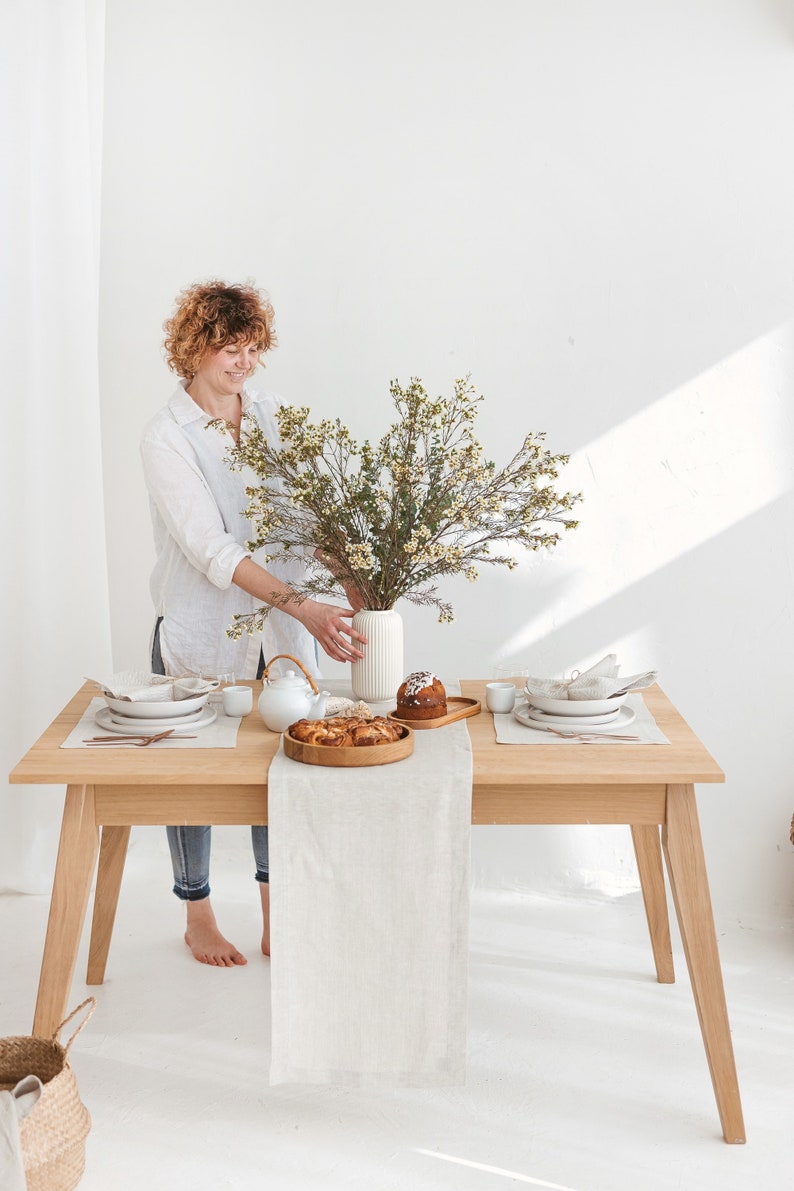 A woman putting a vase with wildflowers on a table decorated with a herri,ngbone white table runner, placemat, and napkin, combined with white plates and copper-colored tableware and wooden serving plates