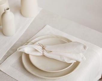 May include: A minimalist table setting featuring two cream-colored plates, a folded linen napkin tied with twine, and a white placemat. Decorative white ceramic vases and a sprig of greenery are in the background, creating a serene, neutral-toned dining scene.