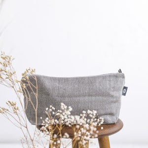 A large grey linen cosmetic bag in a herringbone pattern with a zipper from the side on a stool