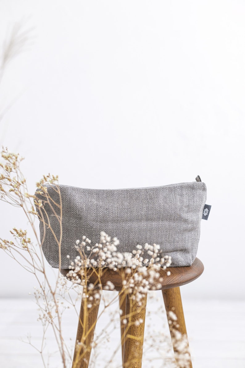 A large grey linen cosmetic bag in a herringbone pattern with a zipper from the side on a stool