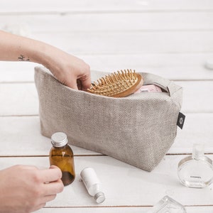 A woman putting a hairbrush in a large white linen cosmetic bag with a zipper decorated in a herringbone pattern