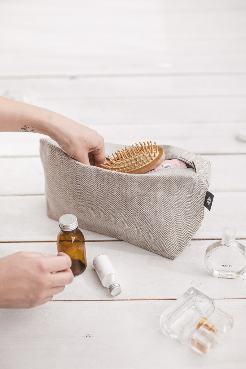 A woman putting a hairbrush in a large white linen cosmetic bag with a zipper decorated in a herringbone pattern