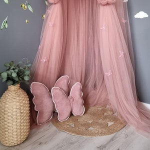 May include: A pink tulle canopy with pom poms and butterfly embellishments. The canopy is set up in a corner of a room with a rainbow wall decal and a wicker vase with greenery.