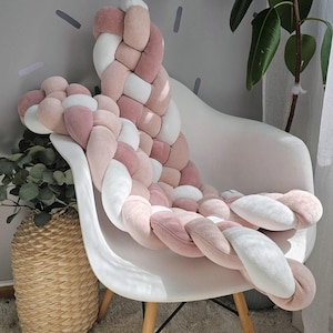 May include: A white chair with wooden legs is adorned with a large, braided cushion in shades of pink and white. The cushion is draped over the back and seat of the chair. A woven basket with greenery sits beside the chair.