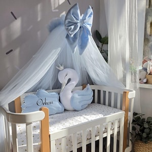 Puede incluir: Una cuna blanca con dosel y lazo azul claro. La cuna tiene un colchón con estampado de corazones dorados, una almohada de cisne y una almohada azul con el nombre "Orhan". La cuna es de madera y tiene un diseño moderno.