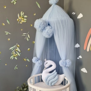 May include: A white and light blue swan-shaped pillow rests inside a white crib. A light blue canopy with pom-poms hangs above the crib. The wall is decorated with olive branch decals and a rainbow.