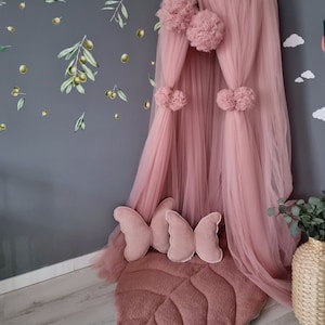 Könnte beinhalten: Ein rosa Tüllbaldachin mit Bommeln hängt über einem rosa blattförmigen Teppich. Zwei rosa Schmetterlingskissen liegen auf dem Teppich.