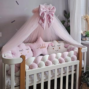 Puede incluir: Una cuna blanca con un protector trenzado rosa y blanco, adornada con almohadas rosas en forma de estrella y nube. Un dosel rosa transparente con un lazo grande está drapeado encima. La ropa de cama de la cuna tiene un estampado de estrellas doradas. La almohada de estrella tiene el nombre "Adele".
