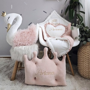 Könnte beinhalten: Ein Kinderzimmer mit einem weißen schwanförmigen Kissen mit goldener Krone, einem rosa kronenförmigen Kissen mit der Aufschrift "Ariana" und einem herzförmigen Kissen mit der Aufschrift "Ariana". Ein weißer Stuhl mit einer Decke und ein Holzregal mit einem Blumenstrauß sind ebenfalls zu sehen.