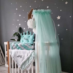May include: A white crib with a mint green tulle canopy and a white and green braided pillow. A mint green pillow with the name "Mark" embroidered on it is on the crib. The crib is decorated with white stars on a gray wall.