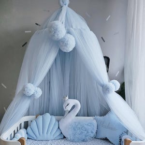 May include: A baby crib with a light blue canopy and pom-poms. The crib is decorated with blue pillows, including a swan-shaped pillow and a star-shaped pillow with text. A braided bumper surrounds the crib.