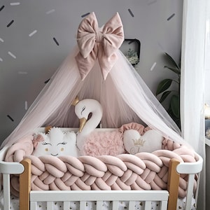 Può includere: Una culla bianca con un paracolpi intrecciato rosa, adornata con cuscini decorativi a forma di nuvola, cuore e cigno. Un baldacchino trasparente con un grande fiocco rosa è drappeggiato sopra la culla, creando un'atmosfera delicata.