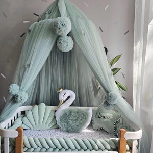 May include: A cot with a mint green canopy, pom-poms, and butterfly details. The cot includes a braided bumper, a swan-shaped cushion, a shell-shaped cushion, and a star-shaped cushion with the name "Eliana".