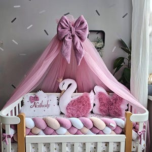 May include: A white crib with a pink canopy and decorative pillows. The canopy is adorned with a large bow. Pillows include a swan and heart shapes. The crib bumper is a braided design in shades of pink, white, and beige.