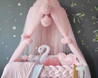 May include: A white crib with a pink braided bumper, a pink tulle canopy with pom-poms, and a pink crochet basket. Inside are a white swan plush with a gold crown, a pink heart-shaped pillow with the word Princess, and a pink cloud pillow.
