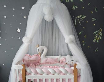 May include: A white wooden crib featuring a braided pink and white bumper, a plush swan with a gold crown, a heart-shaped pillow embroidered with the word Princess, and a cloud pillow, all draped under a sheer white tulle canopy with pom-poms.
