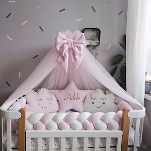 Puede incluir: Una cuna blanca con un parachoques trenzado rosa y blanco, adornada con almohadas rosas en forma de nube y estrella. Un lazo rosa y un dosel transparente coronan la cuna. La almohada de estrella tiene el nombre "Adele".