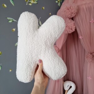 May include: White fluffy letter K pillow. The pillow is being held in a hand in front of a pink tulle canopy with white pom poms.