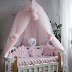 May include: A baby crib with a pink canopy and decorative pillows. The crib features a white frame with a braided bumper and a star-shaped pillow with the name "Cataleya". A swan-shaped pillow and a seashell pillow are also visible.