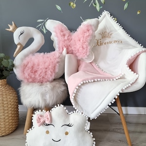 Op de afbeelding: Een wit zwanen knuffel met een roze tule rok, een wit wolkvormig kussen met een roze strik en een wit deken met roze bies en een kroon geborduurd met de naam "Xiomara".