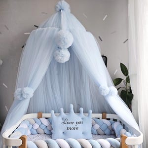 Puede incluir: Una cuna de bebé con dosel azul claro y detalles de pompón. La cuna presenta una almohada en forma de corona con el texto "Love you more". Protectores trenzados en tonos azules y grises rodean la cuna.