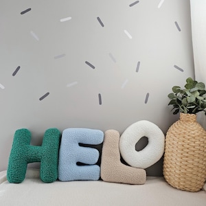 May include: Green, blue, and beige plush letters spelling "HELLO" on a white surface with a gray wall in the background. The wall has gray and white confetti-like shapes.