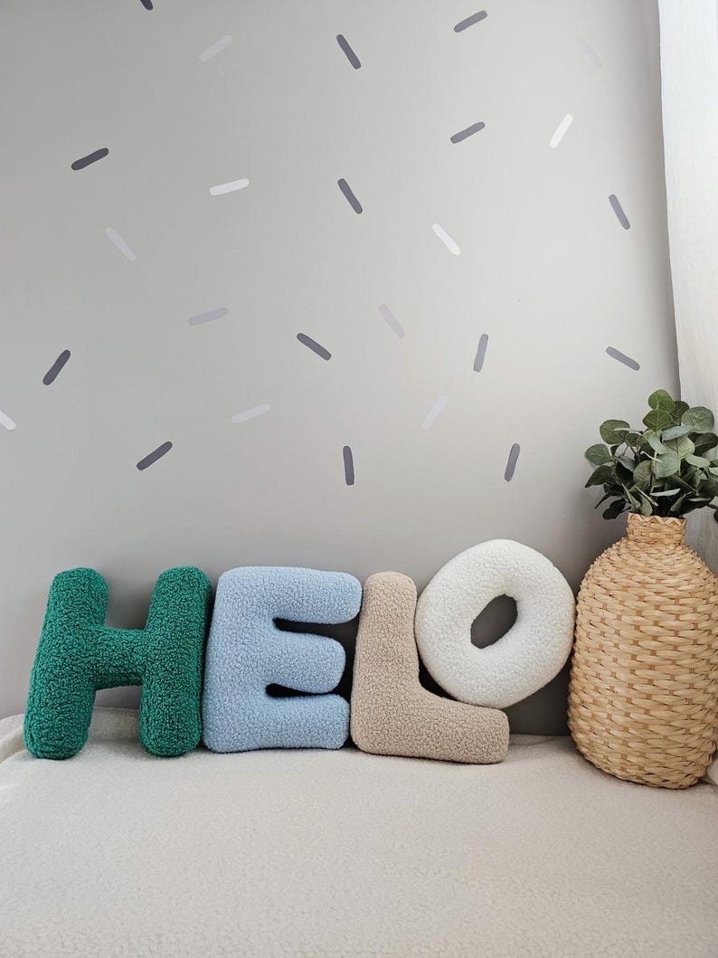 May include: Green, blue, and beige plush letters spelling "HELLO" on a white surface with a gray wall in the background. The wall has gray and white confetti-like shapes.