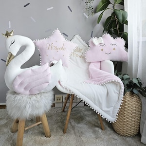 Könnte beinhalten: Ein Kinderzimmer-Szene mit einem weißen Schwan-Plüschtier mit goldener Krone, einem rosa sternförmigen Kissen mit der Aufschrift "Nicole" und einem rosa Wolkenkissen. Eine weiße Decke mit Bommelbesatz ist über einen Stuhl drapiert.