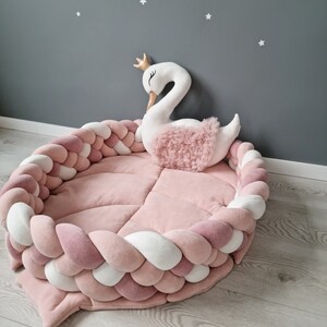 May include: A pink and white braided rope nest with a white swan plush toy with a gold crown on its head. The nest is on a light wood floor.