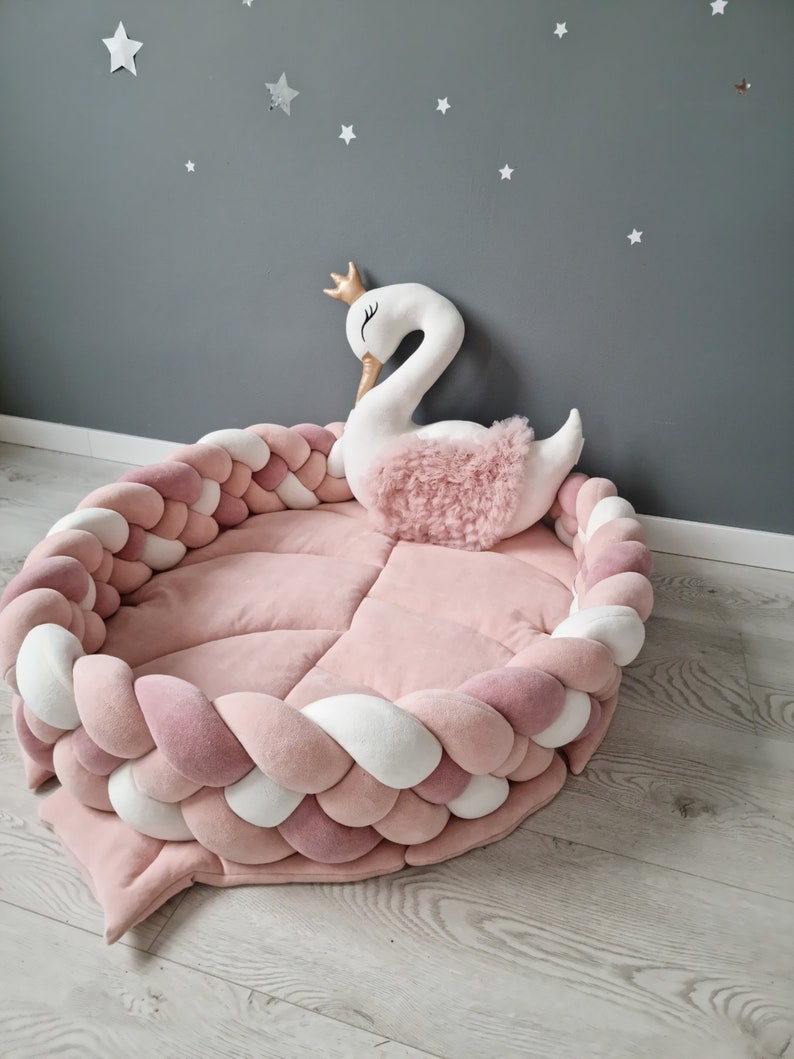 May include: A pink and white braided rope nest with a white swan plush toy with a gold crown on its head. The nest is on a light wood floor.