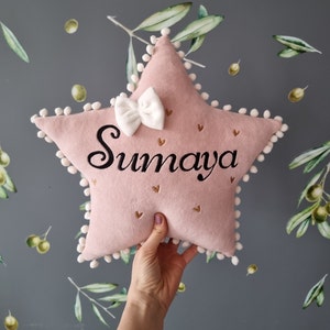 Puede incluir: Una almohada en forma de estrella rosa con pompones blancos alrededor del borde. La almohada tiene un lazo blanco y el nombre "Sumaya" bordado en negro.