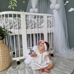 Pu&ograve; includere: Una bambina seduta su un tappetino da gioco a forma di foglia grigio e rosa. Indossa un vestito bianco e una fascia rosa con un fiocco. La bambina &egrave; seduta davanti a un lettino bianco con una zanzariera bianca e paraurti intrecciati grigi e bianchi.