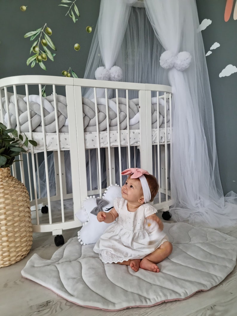 Pu&ograve; includere: Una bambina seduta su un tappetino da gioco a forma di foglia grigio e rosa. Indossa un vestito bianco e una fascia rosa con un fiocco. La bambina &egrave; seduta davanti a un lettino bianco con una zanzariera bianca e paraurti intrecciati grigi e bianchi.