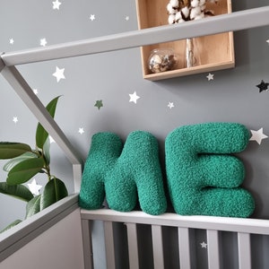 May include: Two large, green, plush letters spelling "ME" are placed on a white crib railing. The letters are made of a soft, fuzzy material. The crib is in front of a gray wall with white star stickers.