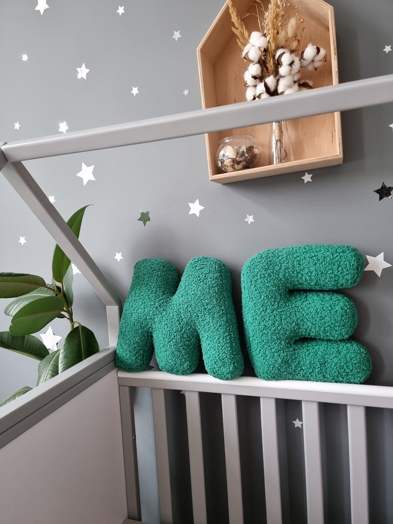 May include: Two large, green, plush letters spelling "ME" are placed on a white crib railing. The letters are made of a soft, fuzzy material. The crib is in front of a gray wall with white star stickers.