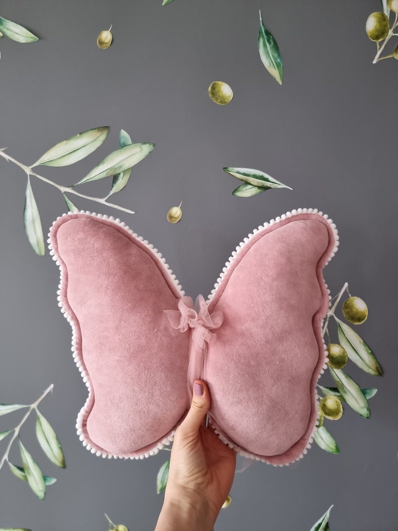 K&ouml;nnte beinhalten: Ein rosa Samt-Schmetterlingskissen mit wei&szlig;em Besatz und einer Schleife aus T&uuml;ll in der Mitte.