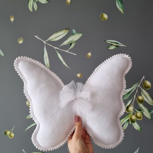 May include: White butterfly-shaped pillow with a white tulle bow in the center. The pillow has a white pom-pom trim around the edges.