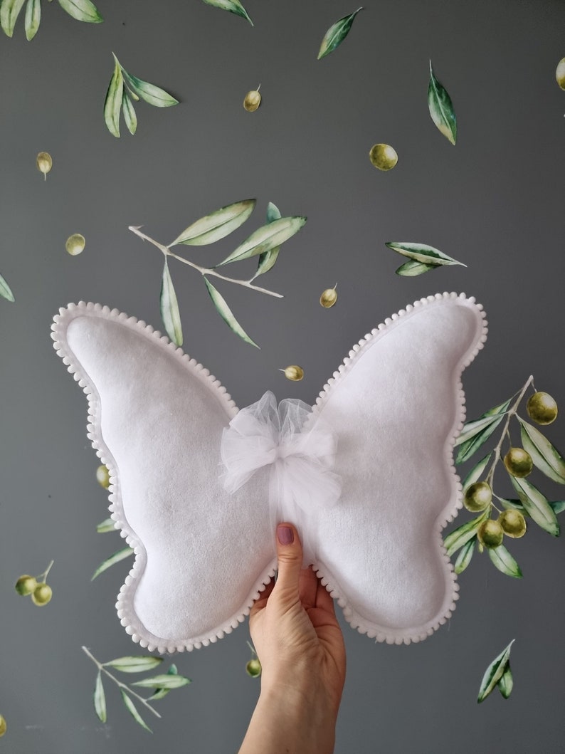 May include: White butterfly-shaped pillow with a white tulle bow in the center. The pillow has a white pom-pom trim around the edges.