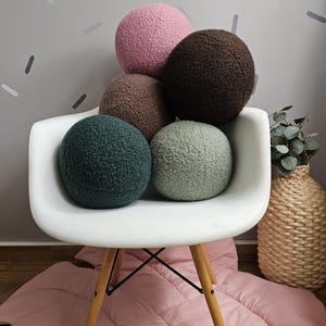 May include: Five round, plush pillows in various colors, including pink, brown, and green, are stacked on a white chair with wooden legs. The chair is on a pink rug.