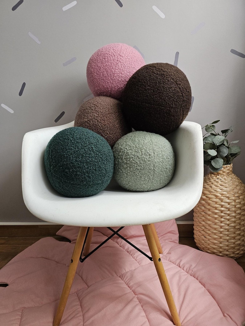 May include: Five round, plush pillows in various colors, including pink, brown, and green, are stacked on a white chair with wooden legs. The chair is on a pink rug.