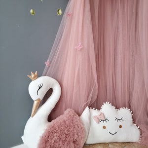 May include: A white swan-shaped pillow with a pink tulle ruffle and a gold crown.  A white cloud-shaped pillow with a pink bow and embroidered eyes and a smile.  Both pillows are on a woven rug.