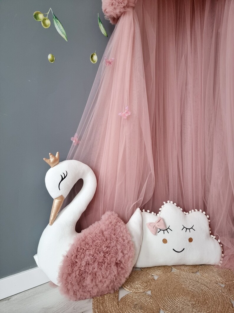 May include: A white swan-shaped pillow with a pink tulle ruffle and a gold crown.  A white cloud-shaped pillow with a pink bow and embroidered eyes and a smile.  Both pillows are on a woven rug.