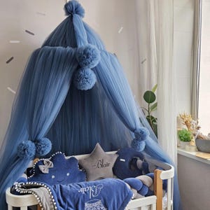May include: A baby crib with a blue tulle canopy adorned with pom-poms. The crib is decorated with star and moon-shaped pillows, a blanket, and a braided bumper. The blanket and star pillow have the name "Blair" embroidered on them.